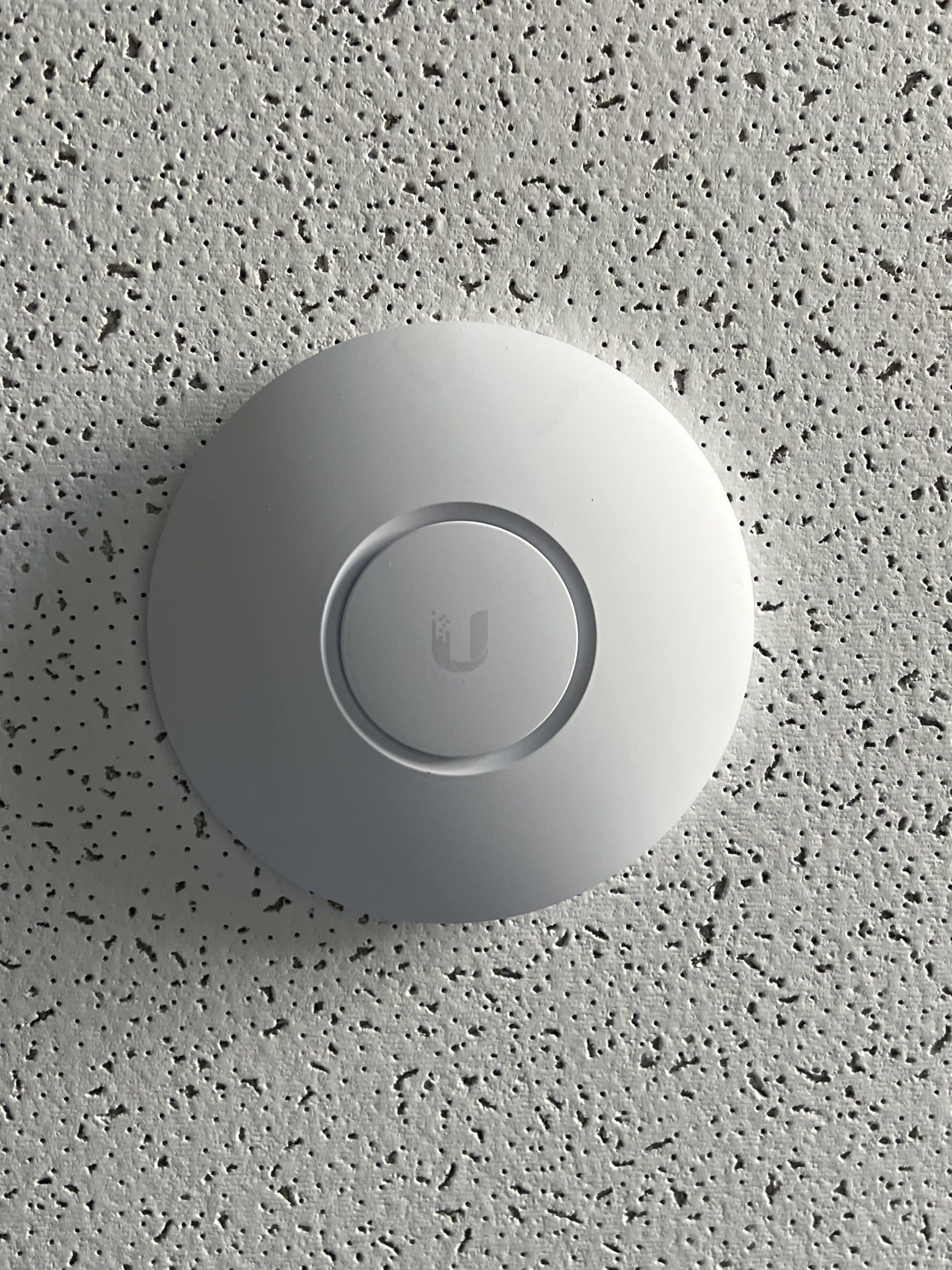 Ubiquiti UniFi Access Point installed in ceiling. White disk shaped with a small circle in the center lit up blue.
